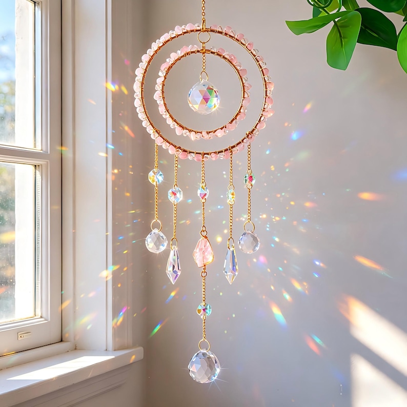May include: A decorative suncatcher with a double-ring design. The rings are adorned with pink beads and gold-colored metal. Clear crystal pendants and faceted crystals hang from the rings, creating a prism effect. The suncatcher is hanging near a window.