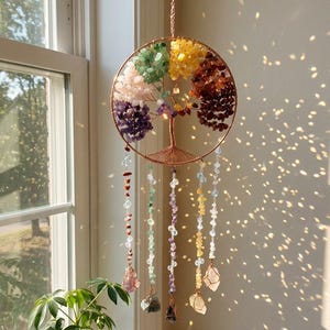Puede incluir: Un colgante de pared de metal color cobre con el árbol de la vida y gemas de colores que representan los chakras. El árbol está suspendido de una cadena y tiene hilos de cuentas con colgantes de gemas.