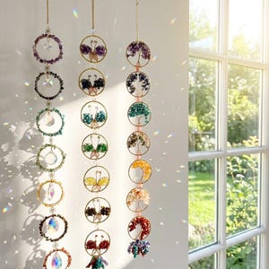 May include: Three hanging crystal suncatchers with gold-colored chains. Each suncatcher features a series of circular designs with colorful gemstones and crystals. Some designs include butterfly and tree motifs. The suncatchers are hanging in front of a window.