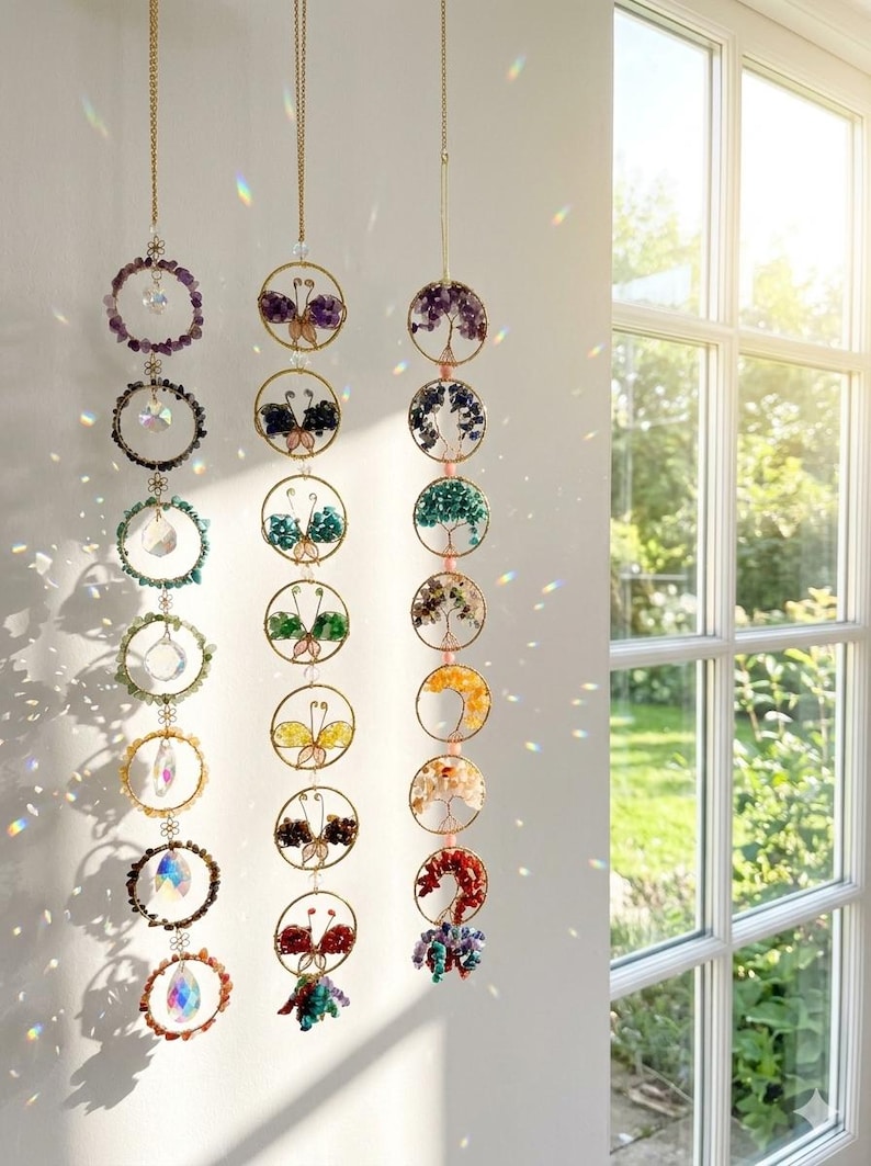 May include: Three hanging crystal suncatchers with gold-colored chains. Each suncatcher features a series of circular designs with colorful gemstones and crystals. Some designs include butterfly and tree motifs. The suncatchers are hanging in front of a window.