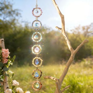 May include: A decorative hanging ornament featuring a series of colorful crystal pendants and circular frames. The frames are adorned with various gemstones and crystals, suspended from a delicate chain. The ornament is displayed outdoors, with a natural backdrop of greenery and sunlight.
