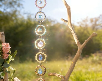 May include: A decorative hanging ornament featuring a series of colorful crystal pendants and circular frames. The frames are adorned with various gemstones and crystals, suspended from a delicate chain. The ornament is displayed outdoors, with a natural backdrop of greenery and sunlight.