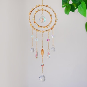 May include: A decorative suncatcher with a gold-colored metal frame. The suncatcher features two concentric circles adorned with small, amber-colored stones. Hanging from the frame are various crystal pendants and spheres, creating a shimmering effect. The overall design is elegant and ethereal.