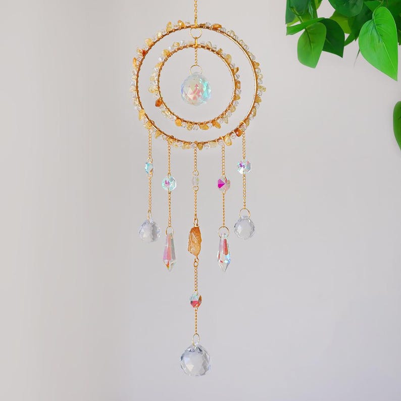 May include: A decorative suncatcher with a gold-colored metal frame. The suncatcher features two concentric circles adorned with small, amber-colored stones. Hanging from the frame are various crystal pendants and spheres, creating a shimmering effect. The overall design is elegant and ethereal.