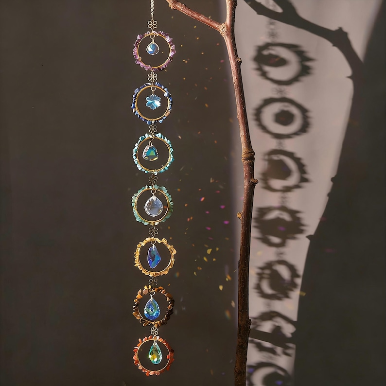 May include: A hanging crystal suncatcher with seven circular frames, each adorned with colorful crystals and a central crystal pendant. The frames are connected by delicate chains, creating a cascading effect. The background is a dark gray with a tree branch.