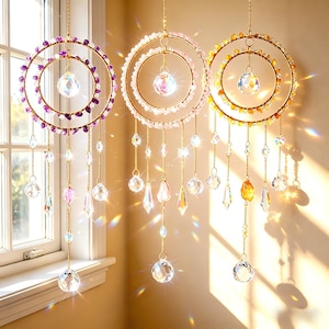 May include: Three hanging crystal suncatchers with gold-colored rings and chains. The rings are adorned with purple, pink, and yellow beads. Each suncatcher has a large crystal ball at the center and dangling crystal pendants.
