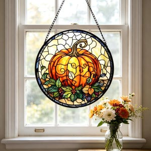 May include: Circular stained glass window depicting a large orange pumpkin with green foliage and berries. The artwork is framed in black and suspended by a chain. A vase of flowers is in front of the window, adding a touch of colour to the scene.