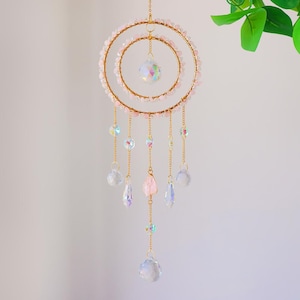 May include: A decorative hanging ornament featuring two gold-colored rings adorned with pink beads. Clear and iridescent crystal beads and teardrop-shaped pendants dangle from gold chains. A large crystal ball hangs in the center, with a smaller one suspended below.