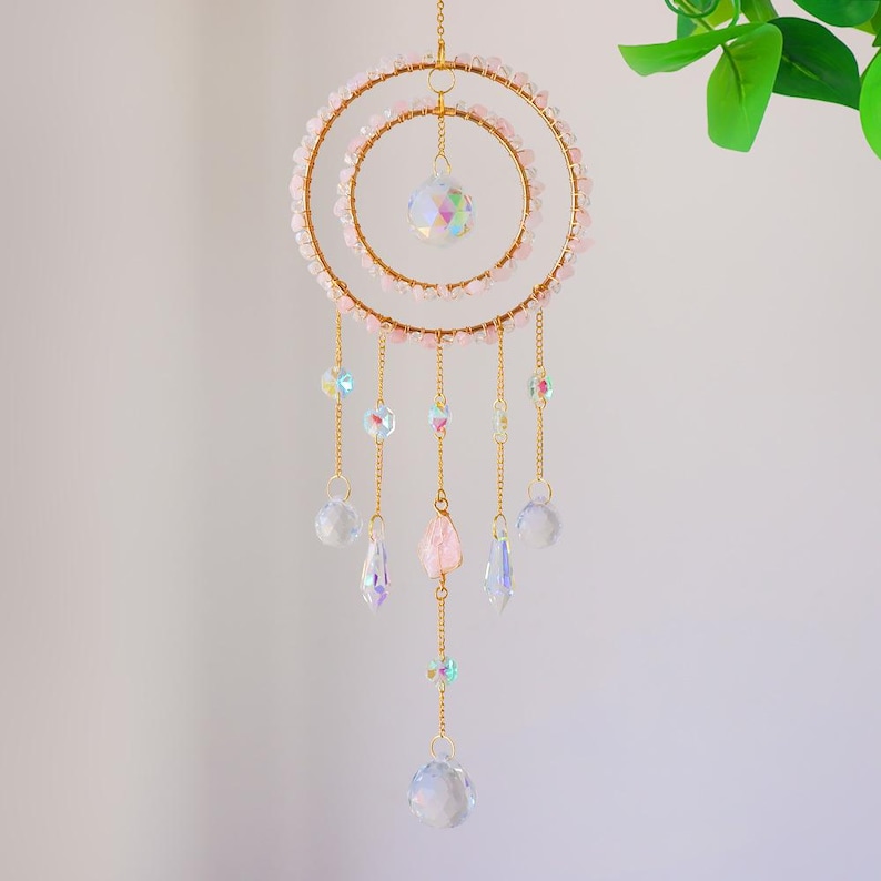 May include: A decorative hanging ornament featuring two gold-colored rings adorned with pink beads. Clear and iridescent crystal beads and teardrop-shaped pendants dangle from gold chains. A large crystal ball hangs in the center, with a smaller one suspended below.