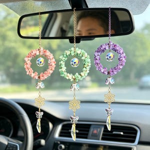Op de afbeelding: Drie auto-ornamenten hangen aan een achteruitkijkspiegel. Elk ornament heeft een cirkelvormige krans van roze, groene en paarse stenen, met een kristallen bol in het midden. Daaronder heeft elk ornament een gouden bloem, vlinder en kristallen hanger.