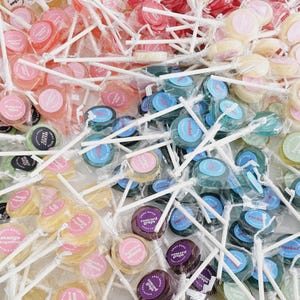 May include: A selection of colourful lollipops in clear wrappers with white sticks. The lollipops are in shades of pink, green, blue, yellow, and purple. Each lollipop has a circular label with text.