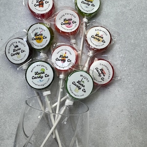 May include: A collection of nine round, individually wrapped lollipops in various colors, each featuring a Luna Candy Co. label with specific cocktail names like Fireball Cinnamon, Apple-Tini, Tipsy Paradise, Island Oasis, Amaretto, Blackberry Mojito, Secret Cocktail, Melon Margarita, and Cosmopolitan.