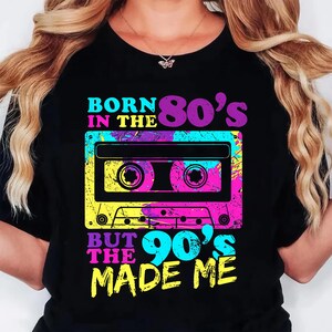 May include: Black t-shirt with a colorful graphic of a cassette tape and text. The text reads "BORN IN THE 80's BUT THE 90's MADE ME" in vibrant colors. The design has a retro, vintage aesthetic.