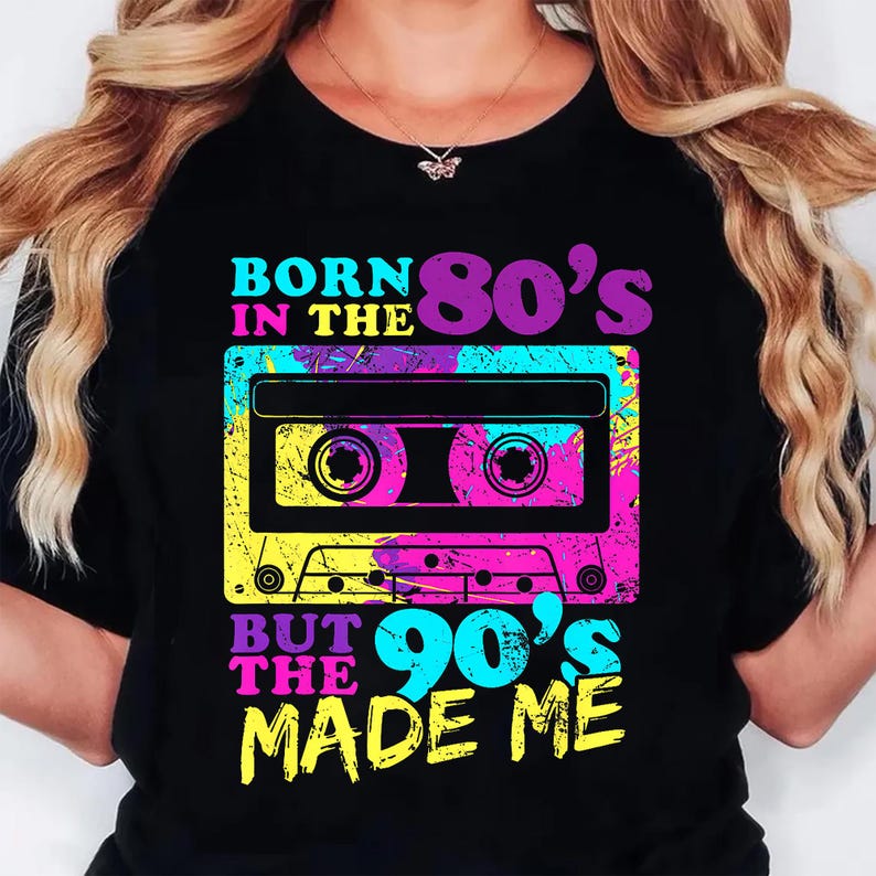 May include: Black t-shirt with a colorful graphic of a cassette tape and text. The text reads "BORN IN THE 80's BUT THE 90's MADE ME" in vibrant colors. The design has a retro, vintage aesthetic.