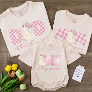 May include: Three matching cream-colored shirts and a onesie with a goose graphic and the words "DAD", "MOM", and "ONE" in pink gingham letters. The text "of the sill" and "silly goose" are also included. A tag reads "With leee MyHeartBeatyShop".