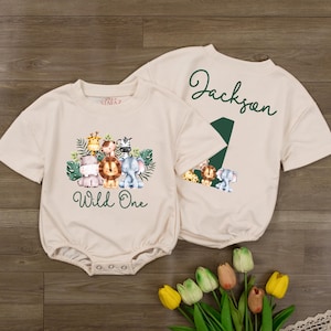 May include: Two beige short-sleeved rompers featuring watercolor jungle animal graphics. One romper displays a group of animals with the text Wild One. The other romper features the name Jackson above a large green number one and animal graphics.