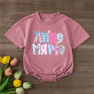 May include: A dusty rose short-sleeved romper featuring the name Anne Marie in colorful, patterned block letters. The text includes floral, striped, and polka-dot designs with small heart and flower accents. The garment has a ribbed crew neckline.