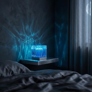 May include: A blue, cube-shaped lamp with a textured surface sits on a shelf. The lamp emits a bright blue light, casting a wave-like pattern on the dark wall. The lamp is a modern home decor item.