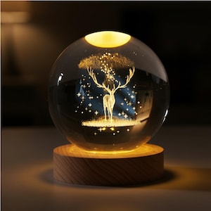 Puede incluir: Una esfera de vidrio transparente con un diseño de ciervo y árbol iluminada por una luz cálida. La esfera se asienta sobre una base de madera redonda, creando una pieza de iluminación decorativa y ambiental. El ciervo y el árbol están grabados en el interior.