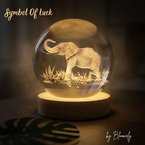 May include: A clear glass sphere with an etched elephant design, illuminated from below by a warm light. The sphere sits on a wooden base, with the text "Symbol Of Luck" and "by Bloomely" in gold script.