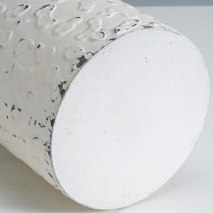 May include: Close-up of a white, cylindrical metal container with a distressed finish. The surface has a textured pattern and areas of black distressing. The bottom is a solid white circle. Ideal for home decor.