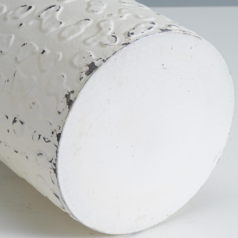 May include: Close-up of a white, cylindrical metal container with a distressed finish. The surface has a textured pattern and areas of black distressing. The bottom is a solid white circle. Ideal for home decor.