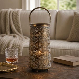 May include: A decorative metal lantern with a handle, featuring a perforated design of circles. The lantern has a warm, antique gold finish and sits on a wooden table. A lit candle inside casts a soft glow. A glass and a tray are also on the table.