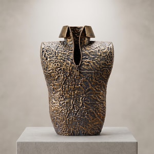 May include: A bronze-toned sculpture shaped like a torso with a textured, crinkled surface. The piece features a structured collar and a central vertical zipper detail that runs down the front, creating a hollow opening in the center of the form.