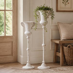 May include: Two tall, white pedestal planters with a distressed finish and ornate handles. The planters feature a classic urn shape supported by slender, tiered stems and circular bases. One planter contains a trailing green plant.