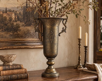 May include: A tall, dark bronze-toned metal urn vase featuring a textured finish, two ornate scroll-shaped handles on the sides, and a central decorative emblem. The vase stands on a pedestal base and contains a branch with small green leaves.