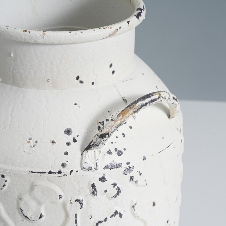 May include: A distressed white metal vase with a handle. The vase has a textured surface with black speckles and a crackled finish, giving it a vintage appearance. The handle is a curved metal bar with a distressed, aged look.
