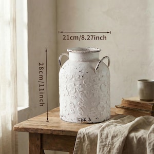 May include: A distressed white metal vase, 11 inches tall and 8.27 inches wide, with two arched handles. The vase has a floral pattern and sits on a wooden table. The vase has a rustic, farmhouse aesthetic.