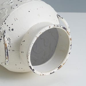 May include: Off-white metal vase with a distressed finish. The vase has two handles and a wide opening. The surface has a textured appearance with dark speckles, giving it a rustic, vintage look. The vase is designed for decorative purposes.