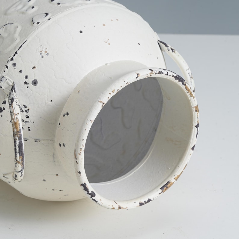 May include: Off-white metal vase with a distressed finish. The vase has two handles and a wide opening. The surface has a textured appearance with dark speckles, giving it a rustic, vintage look. The vase is designed for decorative purposes.