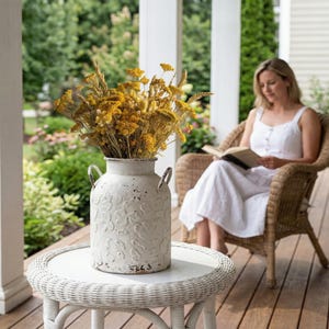 May include: A white, distressed milk jug vase with a floral design, filled with dried yellow flowers. The vase sits on a white wicker table. A woman in a white dress is seated in a wicker chair in the background.