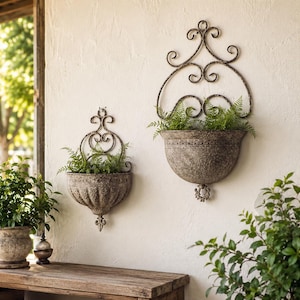May include: Two wall-mounted planters with a weathered, dark metallic finish. Each features a semi-circular basin with a textured surface and an ornate, scrolled metal backplate. Green ferns are planted inside both containers.