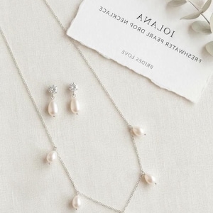 May include: A silver necklace and matching earrings set with freshwater pearl drops. The necklace has multiple pearl pendants, and the earrings feature a single pearl drop. A card with the text "Iolana" and "Brides" is in the background.