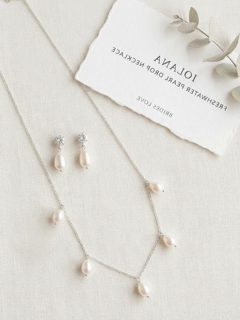 May include: A silver necklace and matching earrings set with freshwater pearl drops. The necklace has multiple pearl pendants, and the earrings feature a single pearl drop. A card with the text "Iolana" and "Brides" is in the background.