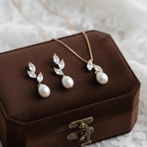 Puede incluir: Un conjunto de joyas dorado con un collar y pendientes, cada uno con una perla en forma de pera y detalles de cristal. El conjunto se exhibe en una caja de joyer&iacute;a de terciopelo marr&oacute;n con un cierre dorado. El fondo es una tela de encaje blanco.