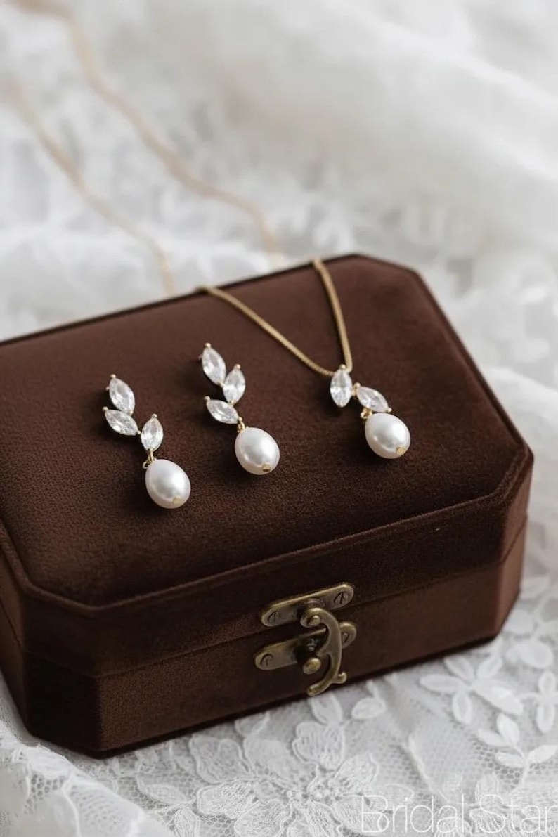 Puede incluir: Un conjunto de joyas dorado con un collar y pendientes, cada uno con una perla en forma de pera y detalles de cristal. El conjunto se exhibe en una caja de joyer&iacute;a de terciopelo marr&oacute;n con un cierre dorado. El fondo es una tela de encaje blanco.