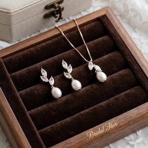 Puede incluir: Un conjunto de joyas dorado con un collar y pendientes, cada uno con un dise&ntilde;o de hojas de cristales transparentes y una perla blanca colgante. El conjunto se exhibe en una caja de madera forrada de terciopelo marr&oacute;n, con las palabras "Bridal Star" en oro.
