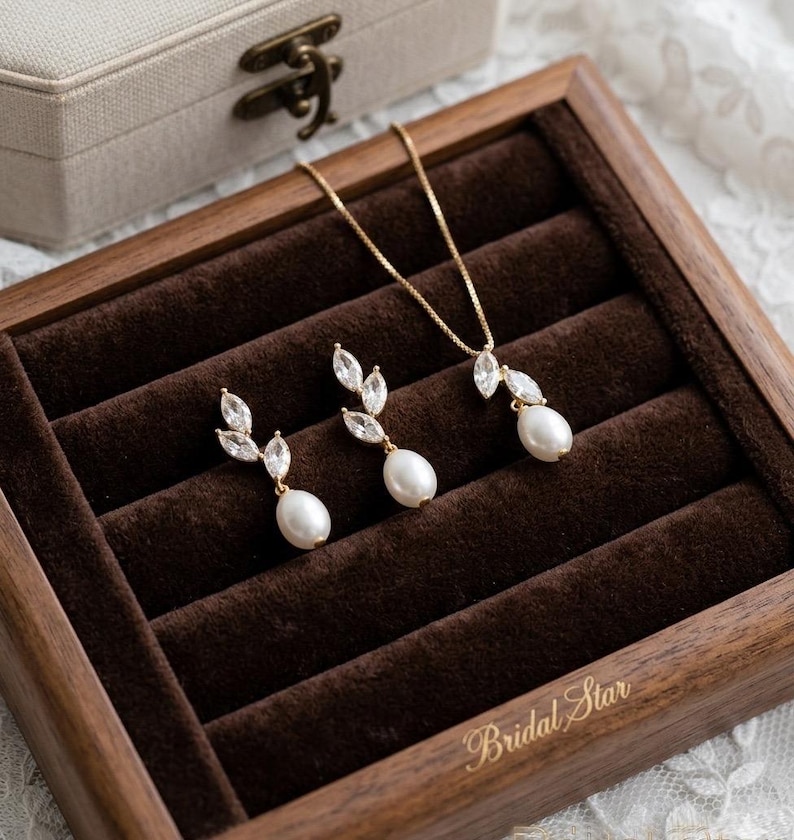 Puede incluir: Un conjunto de joyas dorado con un collar y pendientes, cada uno con un dise&ntilde;o de hojas de cristales transparentes y una perla blanca colgante. El conjunto se exhibe en una caja de madera forrada de terciopelo marr&oacute;n, con las palabras "Bridal Star" en oro.