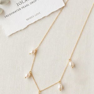 May include: A delicate gold necklace with freshwater pearl drops. The necklace features multiple small, lustrous pearls suspended from a fine gold chain. A card with the text "IOLANA FRESHWATER PEARL DROP NECKLACE BRIDES LOVE" is in the background.