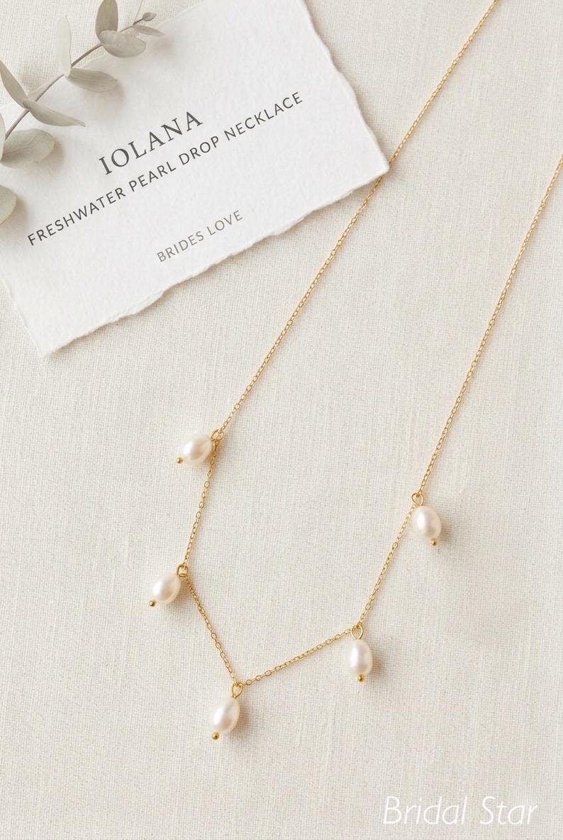 May include: A delicate gold necklace with freshwater pearl drops. The necklace features multiple small, lustrous pearls suspended from a fine gold chain. A card with the text "IOLANA FRESHWATER PEARL DROP NECKLACE BRIDES LOVE" is in the background.