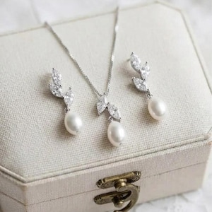 Puede incluir: Un conjunto de joyas plateadas con collar y pendientes, cada uno adornado con cristales transparentes y una perla blanca colgante. El conjunto se exhibe en una caja de joyer&iacute;a beige con un cierre decorativo. El texto "Bridal Star" es visible.