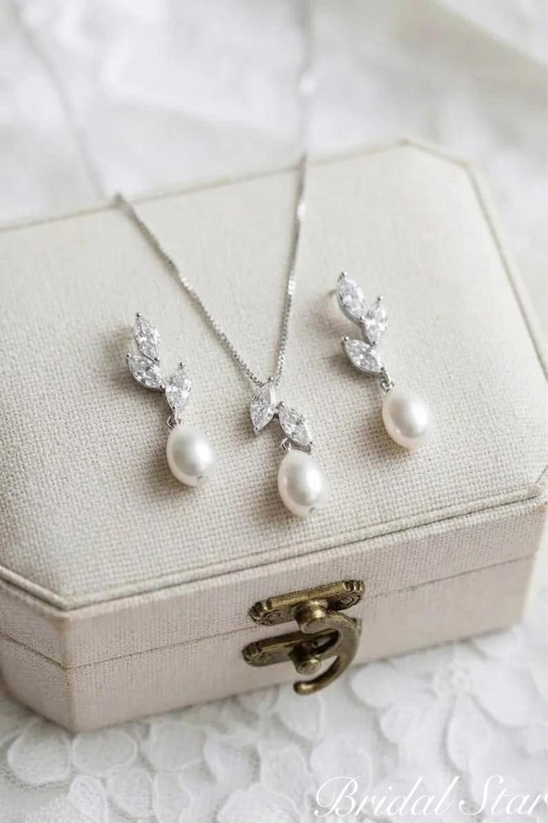 Puede incluir: Un conjunto de joyas plateadas con collar y pendientes, cada uno adornado con cristales transparentes y una perla blanca colgante. El conjunto se exhibe en una caja de joyer&iacute;a beige con un cierre decorativo. El texto "Bridal Star" es visible.