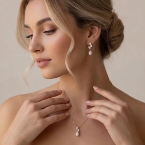 Puede incluir: Un conjunto de collar y pendientes dorados. La joya presenta un colgante de perla en forma de pera y pendientes a juego con detalles de hojas. La mujer tiene el pelo rubio y lleva un top blanco sin tirantes. La imagen est&aacute; bien iluminada.