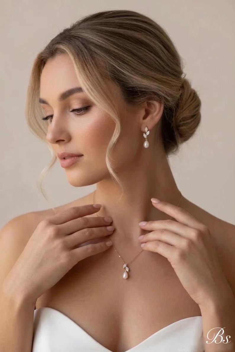 Puede incluir: Un conjunto de collar y pendientes dorados. La joya presenta un colgante de perla en forma de pera y pendientes a juego con detalles de hojas. La mujer tiene el pelo rubio y lleva un top blanco sin tirantes. La imagen est&aacute; bien iluminada.
