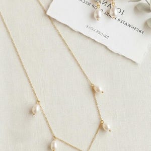 May include: A delicate gold necklace with five dangling pearls, paired with matching pearl earrings. The earrings feature a small diamond-like accent. A card with text is in the background. The jewelry is displayed on a white surface.