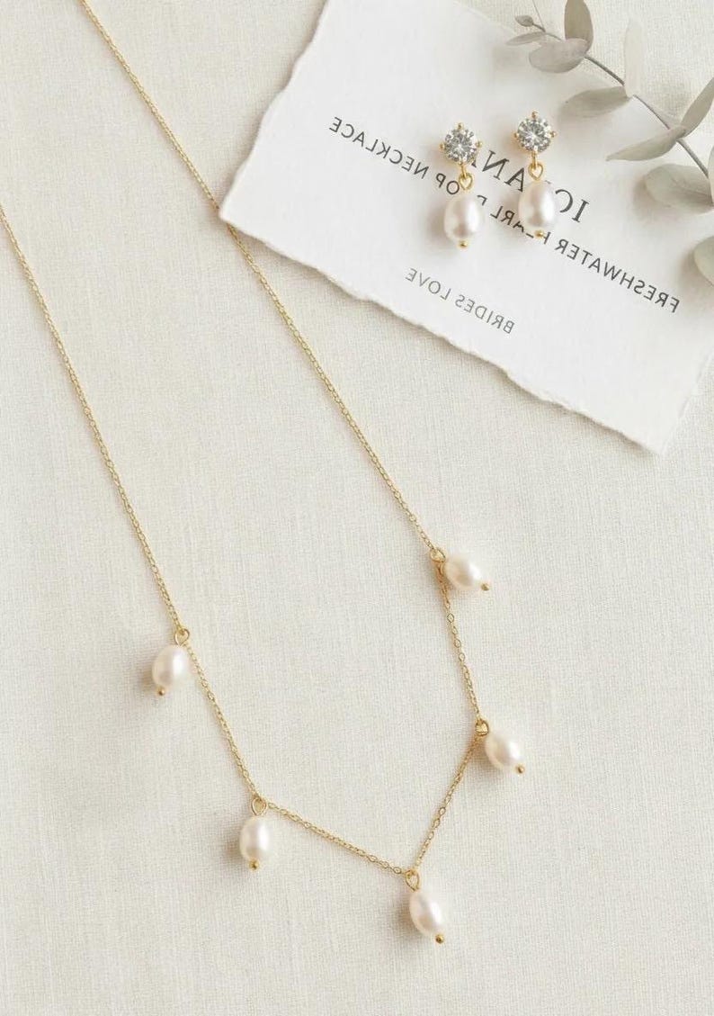May include: A delicate gold necklace with five dangling pearls, paired with matching pearl earrings. The earrings feature a small diamond-like accent. A card with text is in the background. The jewelry is displayed on a white surface.